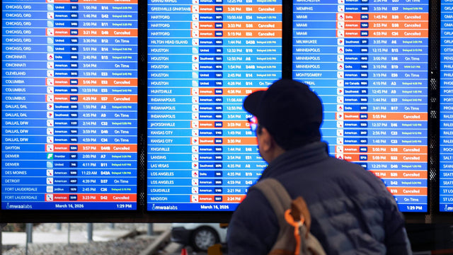 Passengers check a US airport arrivals board showing delayed and canceled flights